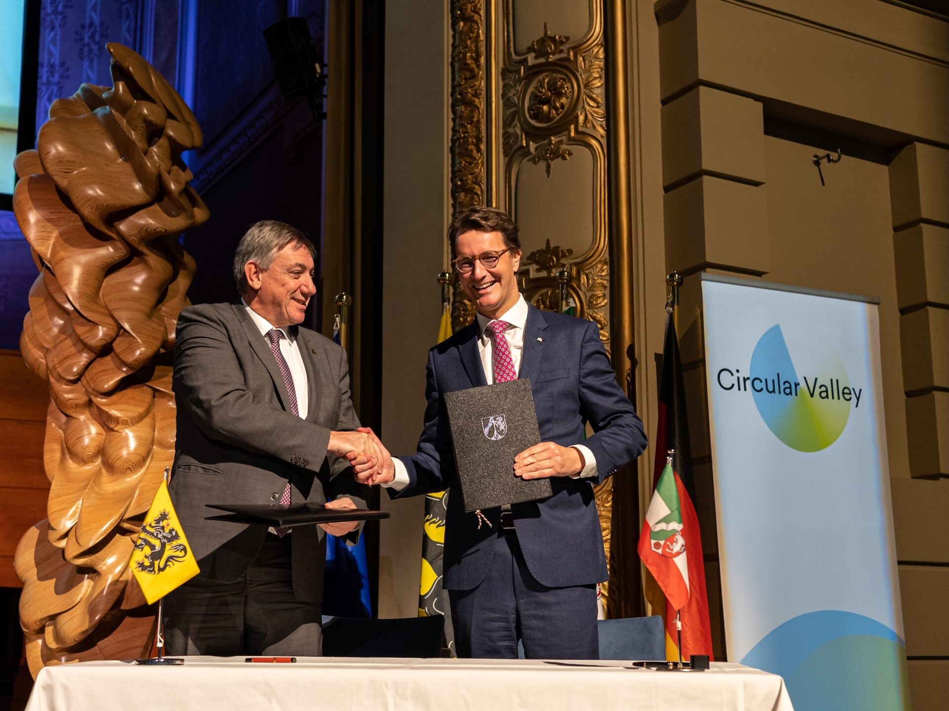 Die Politiker Jan Jambon und Hendrik Wüst reichen sich die Hand und halten ein Dokument, im Hintergrund das Logo von Circular Valley sowie die Flaggen von NRW und Flandern.