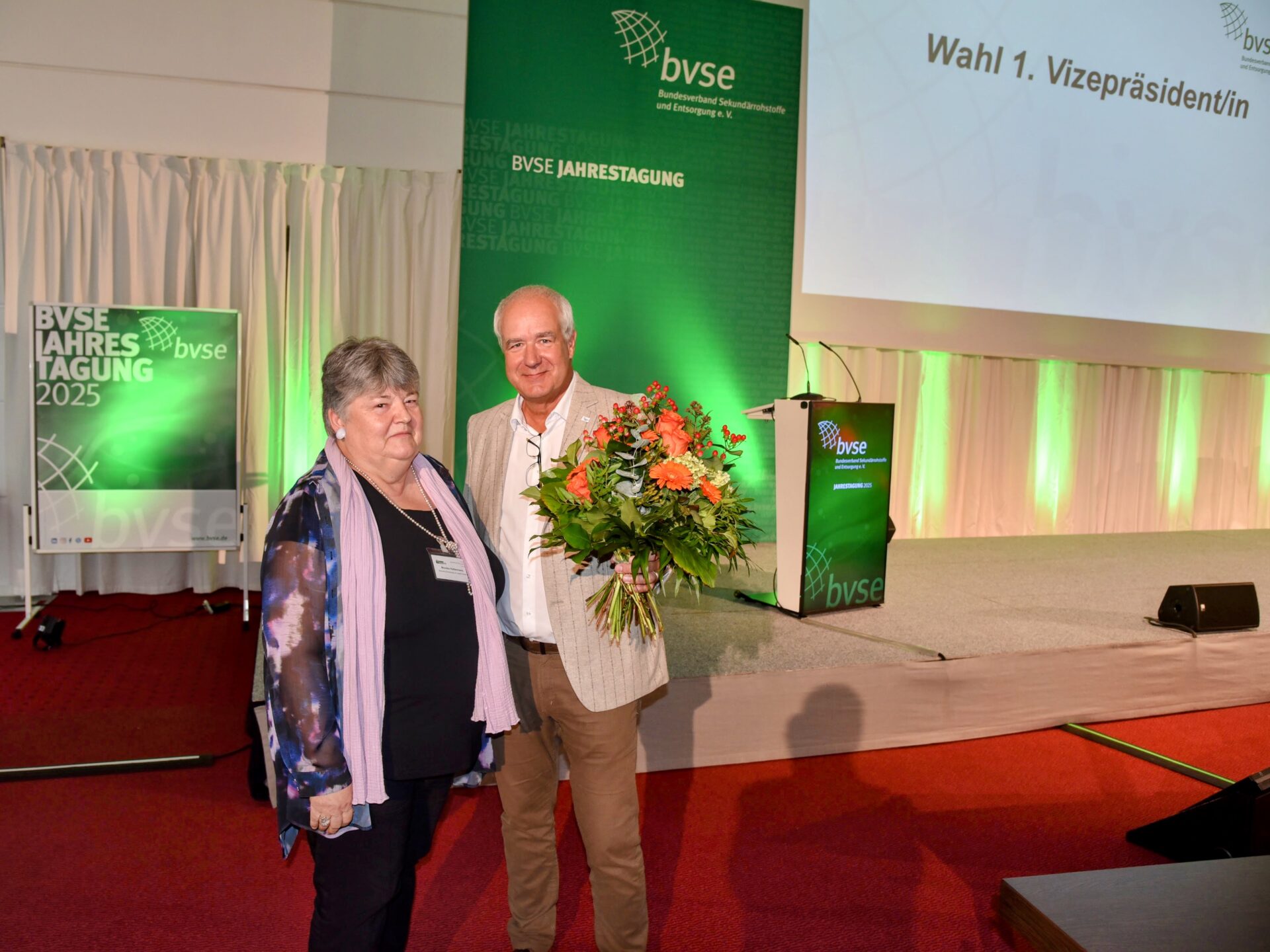 Monika Kattermann steht neben bvse-Präsident Henry Forster, der ihr bei der Jahrestagung des Verbands in Bad Gögging einen Blumenstrauß überreicht. Auf der Leinwand im Hintergrund ist „Wahl 1. Vizepräsidentin“ zu lesen.