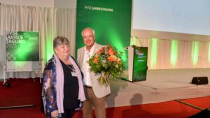 Monika Kattermann steht neben bvse-Präsident Henry Forster, der ihr bei der Jahrestagung des Verbands in Bad Gögging einen Blumenstrauß überreicht. Auf der Leinwand im Hintergrund ist „Wahl 1. Vizepräsidentin“ zu lesen.