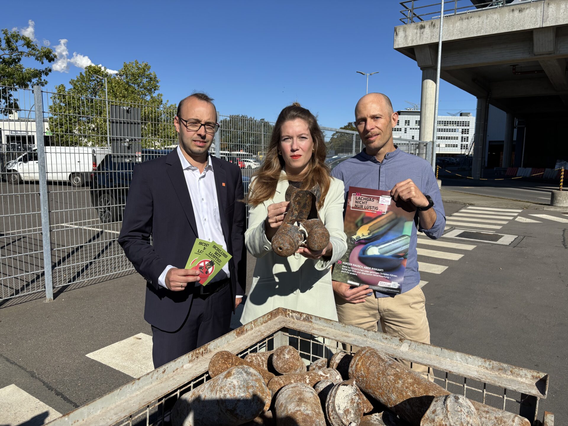 Drei Personen stehen vor einem Container mit entsorgten Lachgas-Kartuschen und halten Info-Materialien sowie eine Kampagnenbroschüre hoch. Aufgenommen vor dem Müllheizkraftwerk Ruhleben.