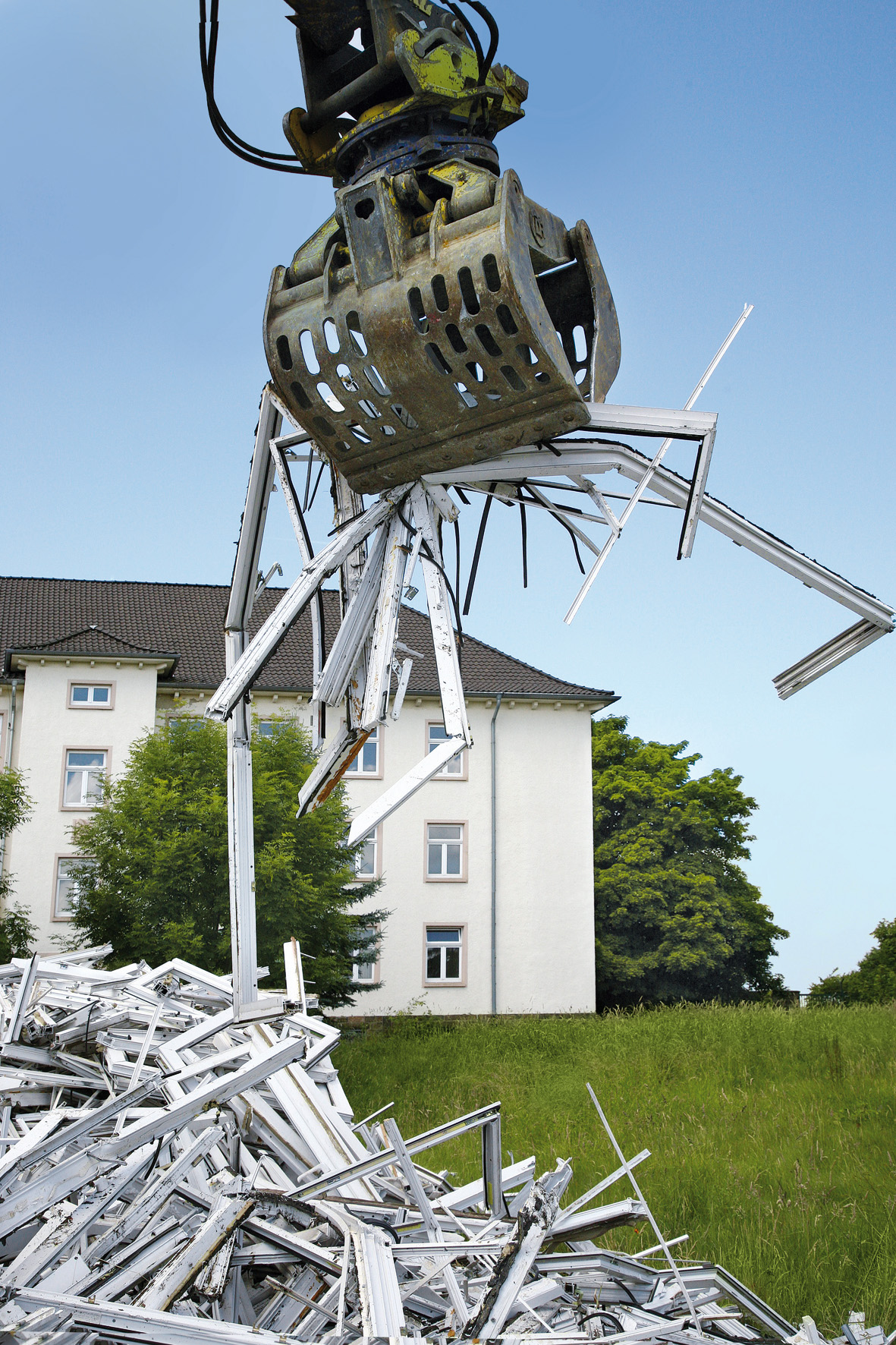 Ein Bagger mit Greifarm hebt alte PVC-Fensterrahmen auf einen Haufen vor einem Gebäude, um sie für das Recycling zu verladen.