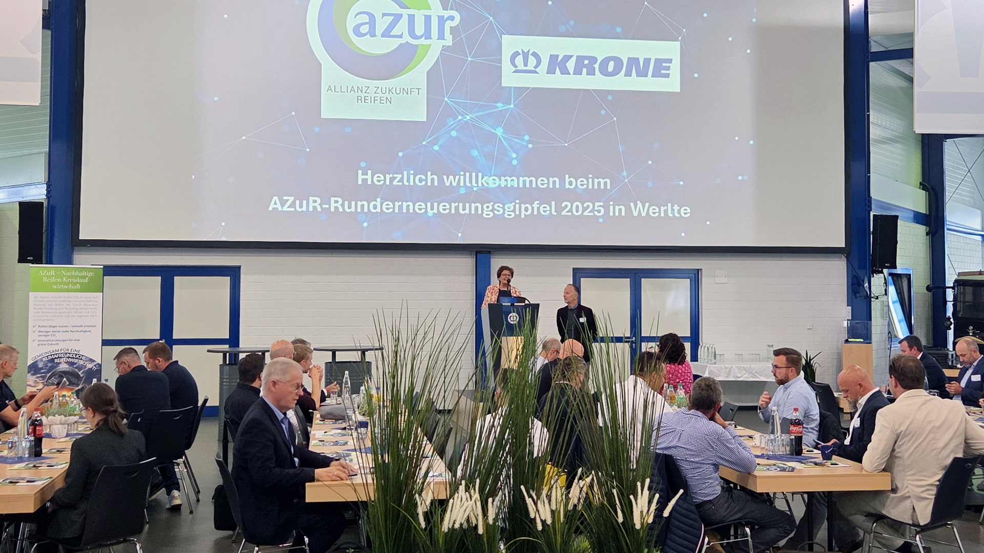 Dr. Frank Albers und Christina Guth stehen auf einer Bühne im Krone-Trailer-Forum und sprechen zur Eröffnung des zweiten Azur-Runderneuerungsgipfels 2025 vor den Teilnehmenden.