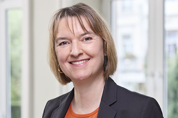 Porträtfoto von Isabell Schmidt, die einen dunklen Blazer trägt und lächelnd vor einem hellen Hintergrund steht